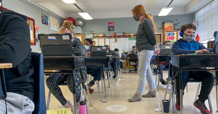teacher walking around to students sitting at desks in classroom