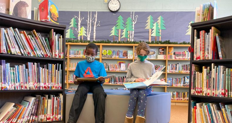 two students reading books in library wearing masks