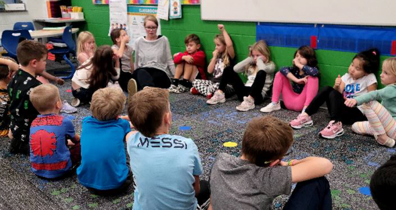 Maple Grove students sit on the floor in a circle with their teacher.