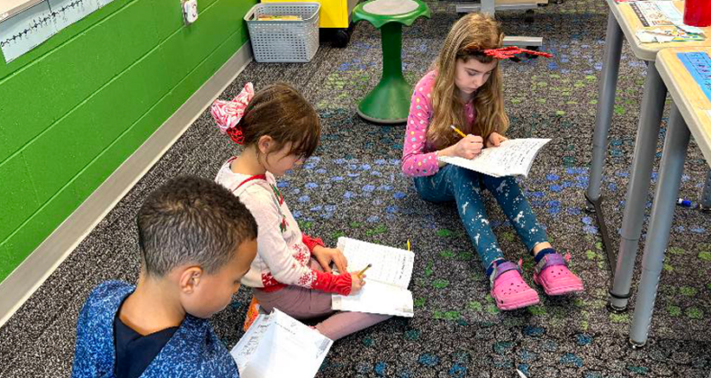 Three Maple Grove students sit on the floor in their classroom while writing in their workbooks.