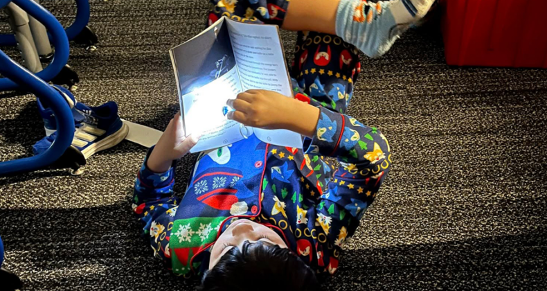 A student lays on the floor with a book in their hand, using a flashlight to read.