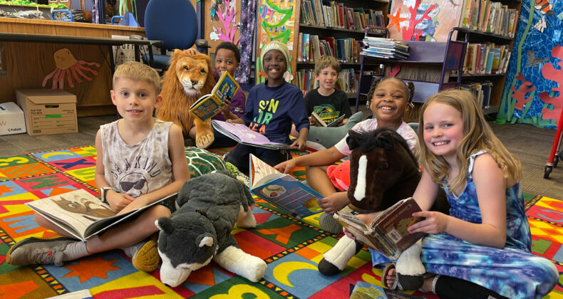 group of student in library sitting down with stuffed animals and books smiling