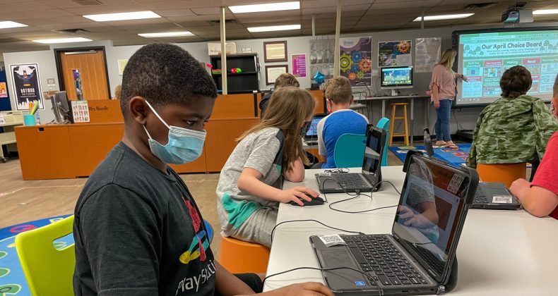 student wearing mask working on laptop at desk in classroom