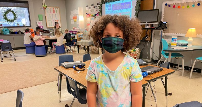 student wearing mask standing in classroom with classmates behind