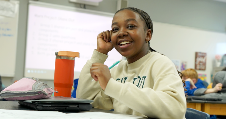 A Harding student smiles at the camera while sitting at a table in a classroom.